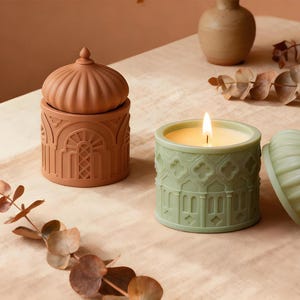 May include: Two decorative candles in terracotta and sage green. The terracotta candle has a domed lid and architectural detailing. The sage green candle is lit, with a flame, and has a similar architectural design. Both candles are on a neutral surface.