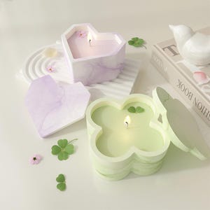 Può includere: Candele a forma di cuore e quadrifoglio con stoppini accesi. Le candele sono in contenitori decorativi con motivi marmorei nei toni del viola e del verde. Piccoli fiori e trifogli sono sparsi intorno alle candele.