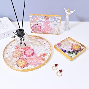 Op de afbeelding: Een verzameling decoratieve harsartikelen met gedroogde bloemen, waaronder een ronde tray, een rechthoekig stuk en een vierkant stuk. Een etherische olie diffuser en hartvormige oorbellen zijn ook aanwezig. De items hebben gouden randen.