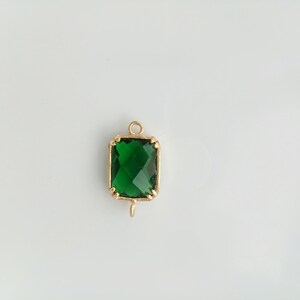 Puede incluir: Un colgante dorado con una piedra preciosa verde cuadrada. El colgante tiene un bucle en la parte superior para sujetarlo a una cadena.