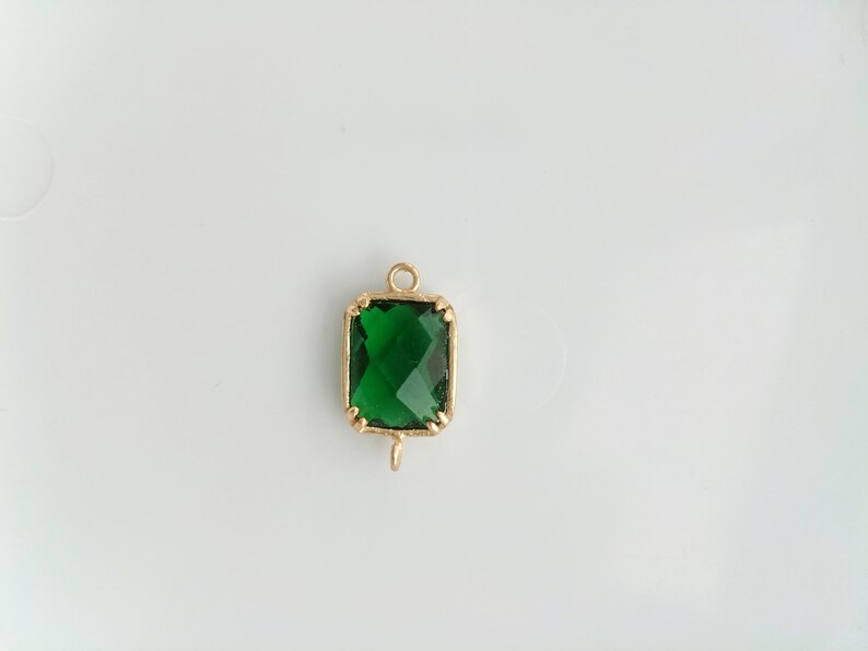 Puede incluir: Un colgante dorado con una piedra preciosa verde cuadrada. El colgante tiene un bucle en la parte superior para sujetarlo a una cadena.