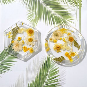 Puede incluir: Dos posavasos de resina transparente con flores secas prensadas. Un posavasos es hexagonal y el otro es redondo. Las flores son amarillas y blancas.