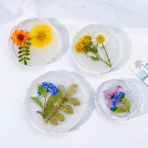 以下が含まれることがあります： 乾燥した花が入った樹脂製のコースター4個セット。各コースターには、オレンジ、黄色、青、紫の花など、さまざまな色の花が透明な樹脂で包まれています。コースターは不規則で有機的なエッジを持っています。