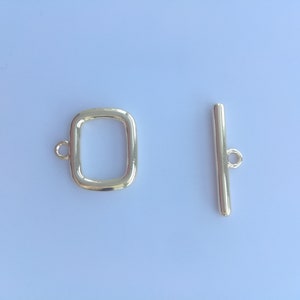 Op de afbeelding: Een gouden metalen schuifgesp met een vierkante lus en een rechte stang. De gesp wordt gebruikt voor kettingen, armbanden of andere sieraden.