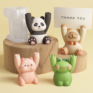 Könnte beinhalten: Eine Sammlung skurriler Tierfiguren, darunter ein Panda, eine Katze, ein Schwein und ein Frosch, alle mit erhobenen Armen. Die Katze hält ein Schild mit der Aufschrift "THANK YOU". Die Figuren sind in verschiedenen Farben gehalten und stehen auf einer Holzoberfläche.
