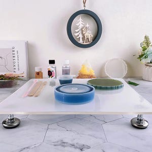 May include: A craft setup with resin art supplies. Includes a blue resin mold, small bottles of liquids, mixing tools, and decorative elements like a tree-shaped candle and a round wall hanging with a deer and tree.