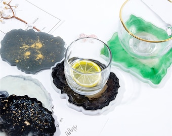 Op de afbeelding: Een verzameling hars coasters in verschillende kleuren, waaronder zwart, groen en wit, met gouden accenten. Een helder glas met een schijfje citroen staat op een zwart-bruine coaster. De coasters hebben onregelmatige, organische vormen.