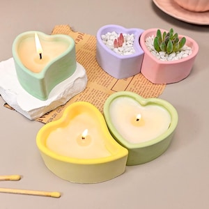 May include: A collection of heart-shaped candles and planters. The candles are in pastel green, yellow, and green containers, with lit wicks. Two planters are filled with white pebbles and small plants, in lavender and pink containers. Matches are visible.