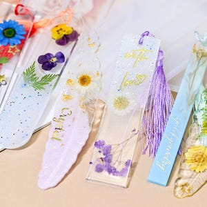 May include: A collection of handmade resin bookmarks with dried flowers and glitter. One bookmark has the words "with love" in gold script. Another has the word "Crystal" in gold script. Each bookmark is a unique piece of art.