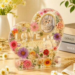 May include: A heart-shaped resin photo frame featuring embedded dried flowers in shades of pink, purple, yellow, and orange. The frame includes the word LOVE, the word Forever, and paw print cutouts. A circular photo of a cat is centred.