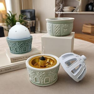 Op de afbeelding: Een verzameling decoratieve wierookbranders in groen en wit. De branders hebben ingewikkelde patronen, met één open om een brandende wierookkegel te laten zien. Stoom stijgt op van de wierook, wat duidt op gebruik. De collectie bevat een koepelvormige brander en een brander met deksel.