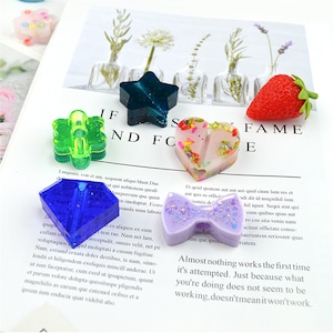 May include: Assortment of colorful resin crafts, including a green four-leaf clover, a blue diamond, a purple bow, a pink heart with glitter, a green star, and a red strawberry. The crafts are on a white surface with text that reads "IF ND FC FAME E".