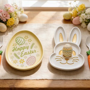 Könnte beinhalten: Zwei Keramikplatten zum Thema Ostern. Eine ist eiförmig und gelb, mit dem Text "Frohe Ostern" und Eiergrafiken. Die andere ist eine weiße, hasenförmige Platte mit goldenen Akzenten, die ein dekoriertes Ei hält. Blumen- und Karottendekorationen sind ebenfalls vorhanden.