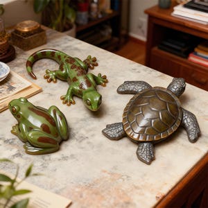 May include: Three ceramic animal figurines: a green frog with brown markings, a green lizard with brown spots, and a bronze-coloured turtle. The figurines are displayed on a marble-patterned surface.