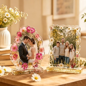 May include: Two clear resin photo frames with embedded pressed flowers. The left frame is oval-shaped with pink and white flowers, holding a wedding photo. The right frame is rectangular with green leaves and white flowers, holding a family photo.