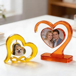 May include: Two heart-shaped photo frames. One is yellow with a photo of a dog, and the other is orange with a photo of a couple. Both frames have a clear, glossy finish and are designed to stand upright on a flat surface.