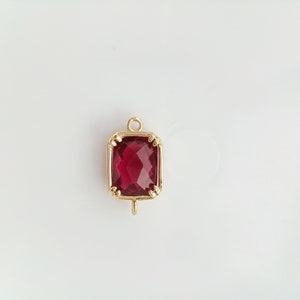 Puede incluir: Un colgante dorado con una piedra preciosa roja en un engaste cuadrado. El colgante tiene un bucle en la parte superior para sujetarlo a una cadena.