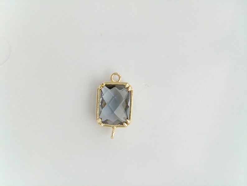 Puede incluir: Un colgante dorado con una piedra preciosa cuadrada transparente y facetada. El colgante tiene un bucle en la parte inferior para sujetarlo a una cadena.