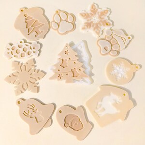 Puede incluir: Una colección de moldes de silicona beige y blancos con temática navideña. Los diseños incluyen un árbol de Navidad, un copo de nieve, una huella de pata, caramelos, una campana con un reno, un guante de Papá Noel y un adorno navideño.