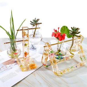 May include: Three clear glass vases with gold accents are displayed on two different resin stands. The stands are decorated with dried flowers and gold leaf. The vases are filled with water and greenery.
