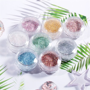 May include: Eight small round jars filled with different coloured glitter. The jars are clear plastic and have a silver lid. The glitter is in shades of pink, gold, green, silver, blue, and red.