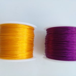 Könnte beinhalten: Zwei Garnrollen, eine gelbe und eine violette, sind auf weißen Kunststoffspulen aufgewickelt.