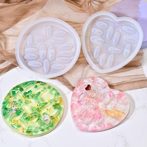 May include: A collection of resin molds in various shapes, including round and heart-shaped designs, each featuring pill-shaped indentations. Two finished resin pieces are also shown, one green and gold, the other pink and white, both heart-shaped.