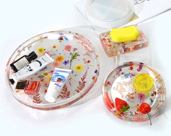 May include: Three clear resin trays with dried flowers and other decorative elements. The largest tray has a round shape and is filled with small flowers and leaves. The medium tray is square and has a bar of soap on top. The smallest tray is round and has a lemon slice, a strawberry, and a cherry on top.