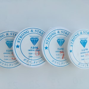 May include: Four spools of white elastic string with blue and red text and a diamond logo. The text on the spools reads "Strong & Stretchy", "Crystal Line", "Water of Crystal", and "Polyurethane 100%".