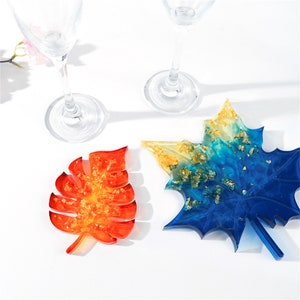 Puede incluir: Tres posavasos de hoja de resina con escamas doradas incrustadas en la resina. Los posavasos tienen forma de hoja de arce, hoja de monstera y hoja genérica. La hoja de arce es azul y amarilla, la hoja de monstera es naranja y amarilla, y la hoja genérica es roja y amarilla.