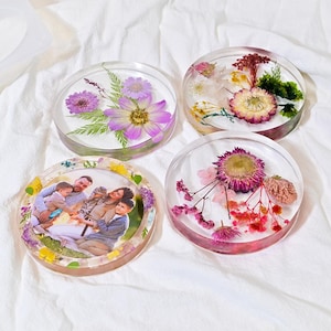 May include: Four round resin coasters. Two coasters feature pressed flowers in shades of purple, yellow, and white. One coaster has a family photo. The fourth coaster has pink and red flowers. All coasters are on a white surface.