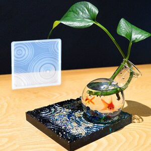 May include: A clear glass vase with a round bottom and a side opening, containing water and two orange goldfish. A green plant with leaves is growing in the vase. The vase is sitting on a black square resin coaster with a blue and gold wave pattern.