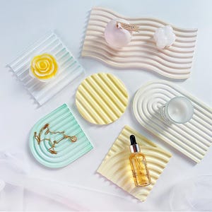 Puede incluir: Una vista cenital muestra una colección de bandejas decorativas de diversas formas y colores. Las bandejas están adornadas con elementos como velas, joyas y un frasco con cuentagotas. La paleta de colores incluye tonos pastel de azul, verde, amarillo y beige, creando una disposición visualmente atractiva.