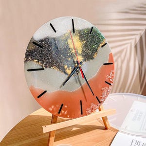 May include: A round decorative clock with a resin face. The face features a blend of colors including orange, gray, black, and gold. The clock is supported by a small wooden easel. The clock hands are black with a red second hand.