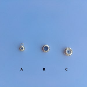 以下が含まれることがあります： 中央に小さな穴が開いた金色の金属ビーズが3つあります。ビーズは左から右に並べてあり、A、B、Cとラベル付けされています。Aとラベル付けされたビーズには、上にループが付いています。ビーズは薄い青色の背景の上にあります。