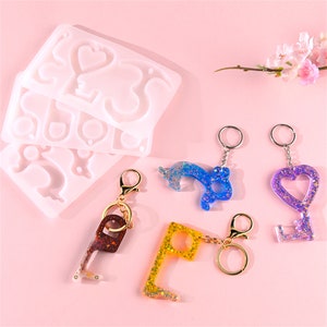 Könnte beinhalten: Ein Satz Silikonformen zum Herstellen von Schlüsselanhängern. Die Formen sind in Form von Herzen, Schlüsseln und anderen Designs. Es gibt vier fertige Schlüsselanhänger aus Glitzerharz in Blau, Lila, Gelb und Braun.