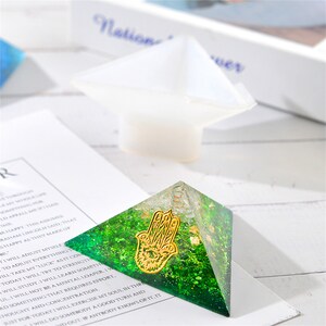 Può includere: Una piramide di resina verde e dorata con un ciondolo a mano di hamsa. La piramide è appoggiata su una superficie bianca.
