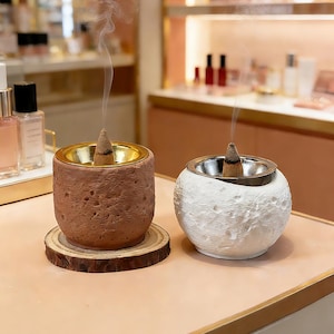 Puede incluir: Dos porta inciensos, uno cilíndrico y otro esférico, cada uno con un cono de incienso encendido. El porta inciensos cilíndrico es marrón con interior dorado, sobre un posavasos de madera. El porta inciensos esférico es blanco con interior plateado.