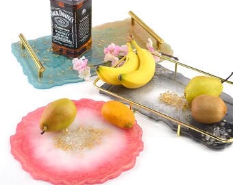 Puede incluir: Tres bandejas de resina con detalles dorados y un asa dorada. Las bandejas son de diferentes colores: azul, gris y rosa. La bandeja azul tiene escamas doradas, la bandeja gris tiene plátanos y la bandeja rosa tiene una pera y un mango.