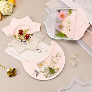 May include: A collection of handmade resin trays in various shapes, including a star, a crescent moon, and a decorative shape. The trays are light pink and contain dried flowers, jewelry, and a card with text. Two pearl earrings are also visible.
