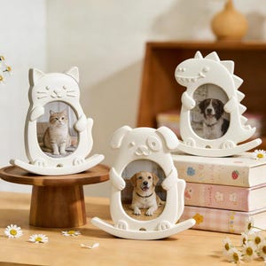 May include: Three white ceramic picture frames in the shapes of a cat, dog, and dinosaur. Each frame holds a photograph of a pet. The cat frame is on a wooden stand, while the dog and dinosaur frames sit on a stack of books.