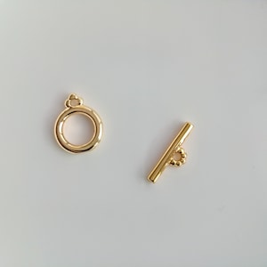 Op de afbeelding: Gouden metalen sluiting met een ronde ring en een rechte stang met een kleine ring aan het ene uiteinde.