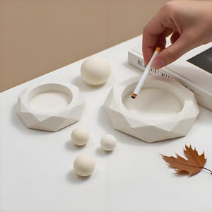 Op de afbeelding: Een verzameling witte, geometrische asbakken en kleine bollen op een wit oppervlak. Een hand houdt een aangestoken sigaret boven een asbak. Een gedroogd blad voegt een vleugje natuurlijke kleur toe.