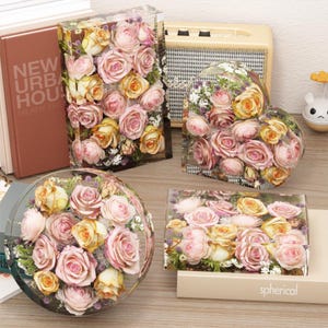 May include: Four decorative floral arrangements encased in clear resin. The arrangements feature pink and yellow roses in rectangular, heart-shaped, spherical, and rectangular forms. The arrangements are displayed on a wooden surface.