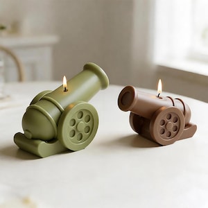 May include: Two cannon-shaped candles. One is olive green, the other is brown. Each candle has a lit wick. The candles are on a light-colored surface. The wheels and barrel details are visible. The candles are decorative.