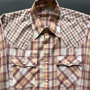Puede incluir: Una camisa vintage a cuadros en crema, beige, rojo y azul marino. La camisa tiene una botonadura, dos bolsillos delanteros con cierre de botón y un cuello puntiagudo. La etiqueta de la marca Levi's es visible.