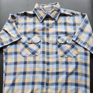 May include: A button-down shirt featuring a plaid design in blue, tan, and brown hues. It has two chest pockets and a traditional collar. The fabric seems to be of a medium weight.