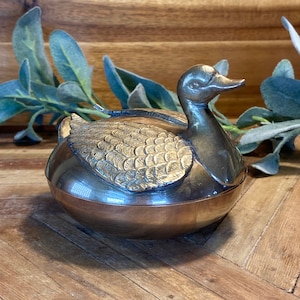 May include: A silver and gold duck-shaped trinket box with a detailed design on the duck's body. The box is sitting on a wooden surface.