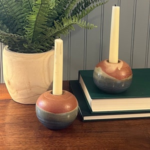 May include: Two ceramic candle holders with a brown, blue, and green glaze. Each holder has a white candle inside. The holders are on a wooden surface with a book in the background.