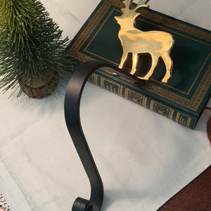 May include: A stocking holder featuring a polished gold-tone metal reindeer silhouette attached to a curved matte black metal hook. The reindeer is standing upright with its head turned, and the hook has a decorative scrolled end at the bottom.